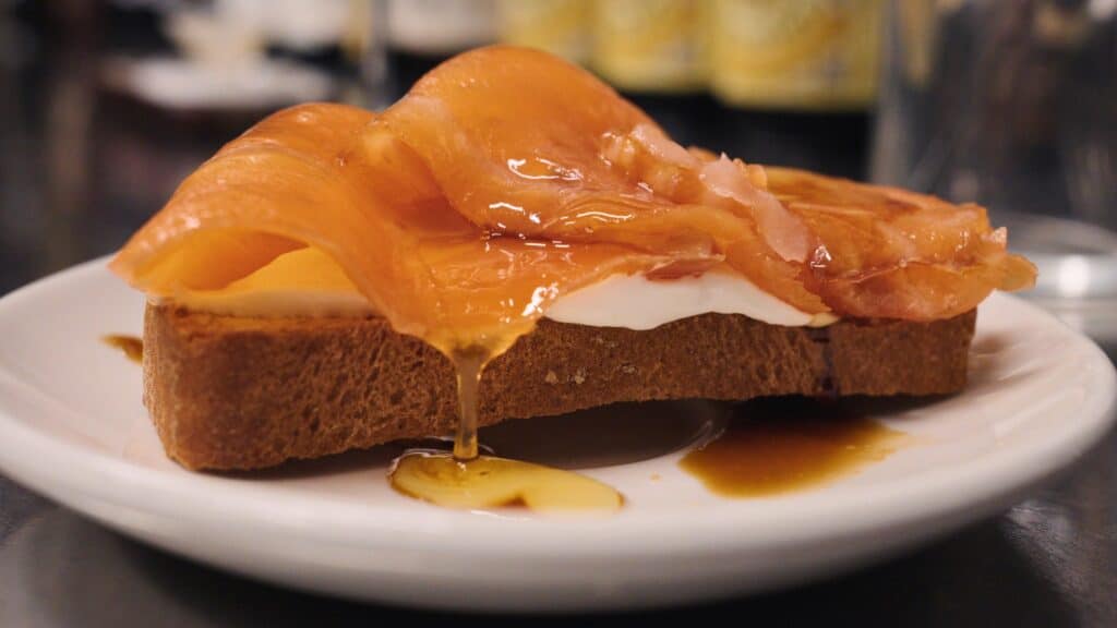 Thick slices of smoked salmon layered over toast with creamy cheese and glossy sauce dripping onto a white plate. Rich seafood dishes like this are among the best food in Barcelona for brunch and tapas lovers.