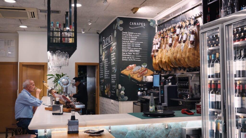 A bright tapas bar with hanging jamón legs, a chalkboard menu labeled "Canapés," and a customer seated at the counter enjoying a drink. The lively setting captures the authentic vibe of restaurants in calle ibiza madrid.