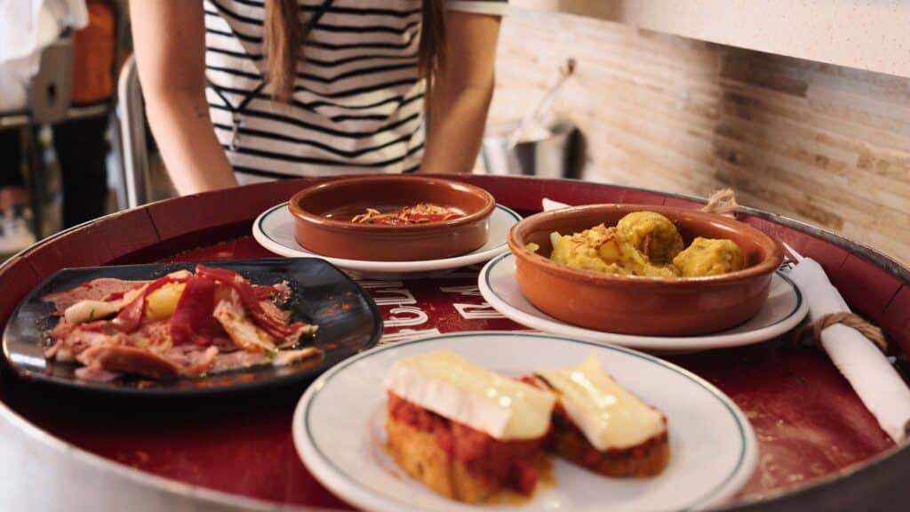 Table filled with Spanish tapas including cured meats, croquettes, and bread topped with melted cheese at a casual restaurant in Madrid. Sharing small plates like these is part of the cost of food in Madrid at traditional tapas bars.