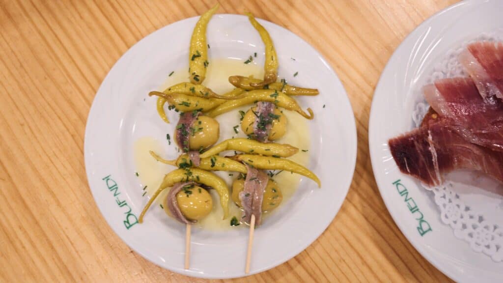 Small plate of skewered olives, anchovies, and yellow peppers drizzled with oil, served alongside slices of cured ham. These traditional tapas are a staple at restaurants in calle ibiza madrid.