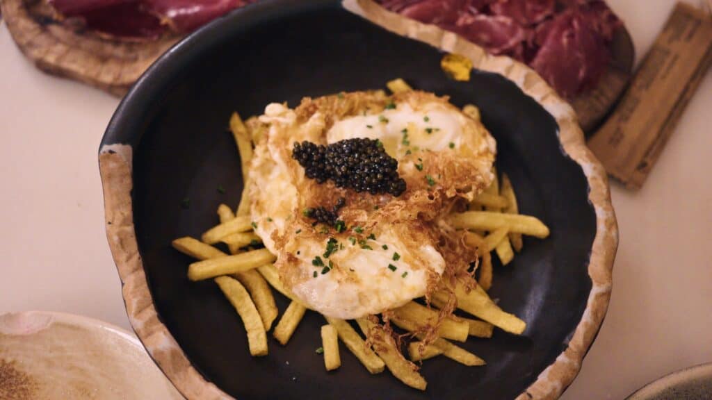 Fried eggs topped with black caviar served over crispy fries in a black bowl with cured meats in the background. Upscale tapas plates like this are featured at some of the best restaurants in Barcelona.