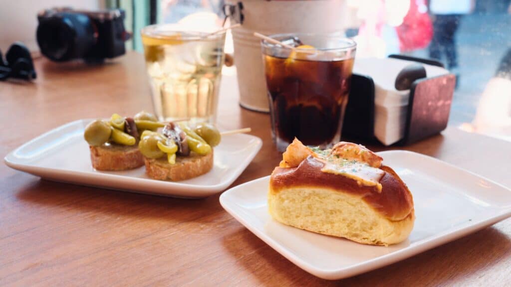 Table spread with small pintxos, olives, peppers, bread rolls, and drinks inside a cozy café in Bilbao. The casual tapas style meal reflects the lively atmosphere of the best restaurants in Bilbao.