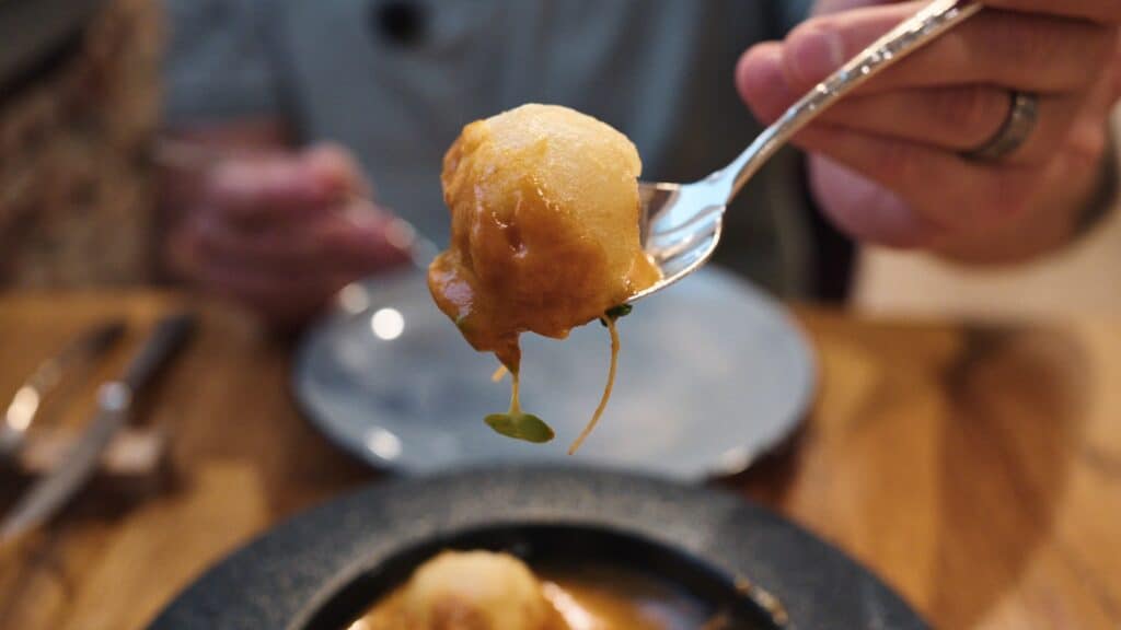 Close up of a fork lifting a bite sized dumpling covered in sauce, with a blurred dining table in the background. The rich texture and plating showcase the comforting dishes served at restaurants in calle ibiza madrid.