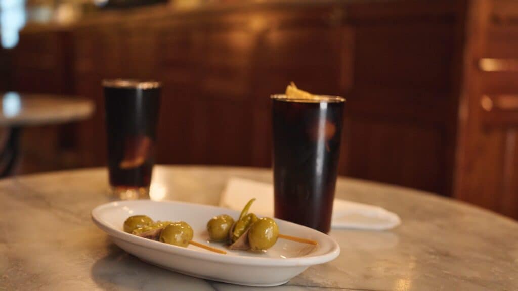Small plate of green olives skewered with anchovies served beside two dark drinks on a marble table. Simple tapas and vermouth pairings are a classic answer to where to eat in Barcelona like a local.