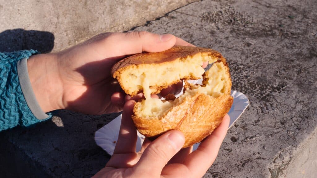 ands pulling apart a warm Pastel Vasco filled with custard while sitting outdoors in sunlight. The gooey filling and crisp crust highlight comforting snacks from the best restaurants in Bilbao.