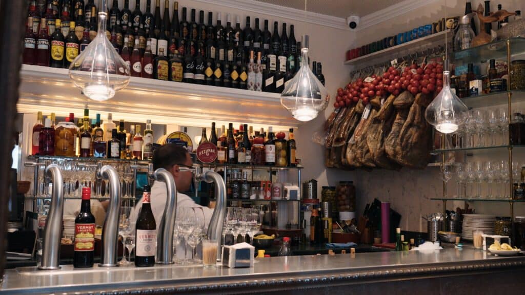 Cozy bar interior lined with shelves of wine and liquor bottles, with cured hams hanging on the wall and a bartender working behind the counter. The warm, bustling atmosphere reflects the classic dining experience at restaurants in calle ibiza madrid.