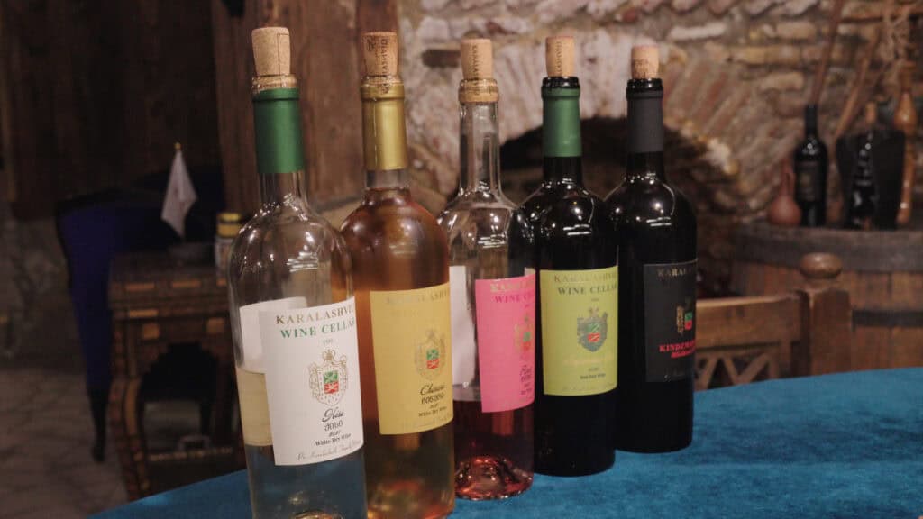 Row of wine bottles with labels reading "Karalashvili Wine Cellar" displayed on a table in a rustic cellar, representing Georgian wine culture alongside traditional Georgian foods.