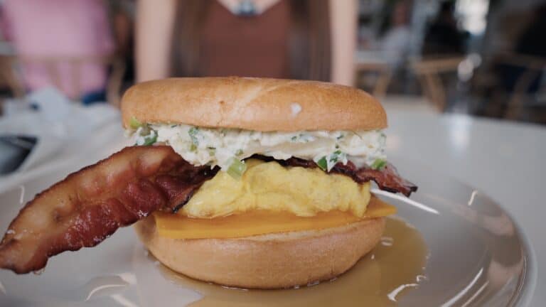 Breakfast sandwich layered with scrambled eggs, cheddar cheese, crispy bacon, and herbed cream cheese on a toasted bun with syrup drizzled on the plate. Creative breakfast dishes make this a standout among the best restaurants in Fort Dodge Iowa.