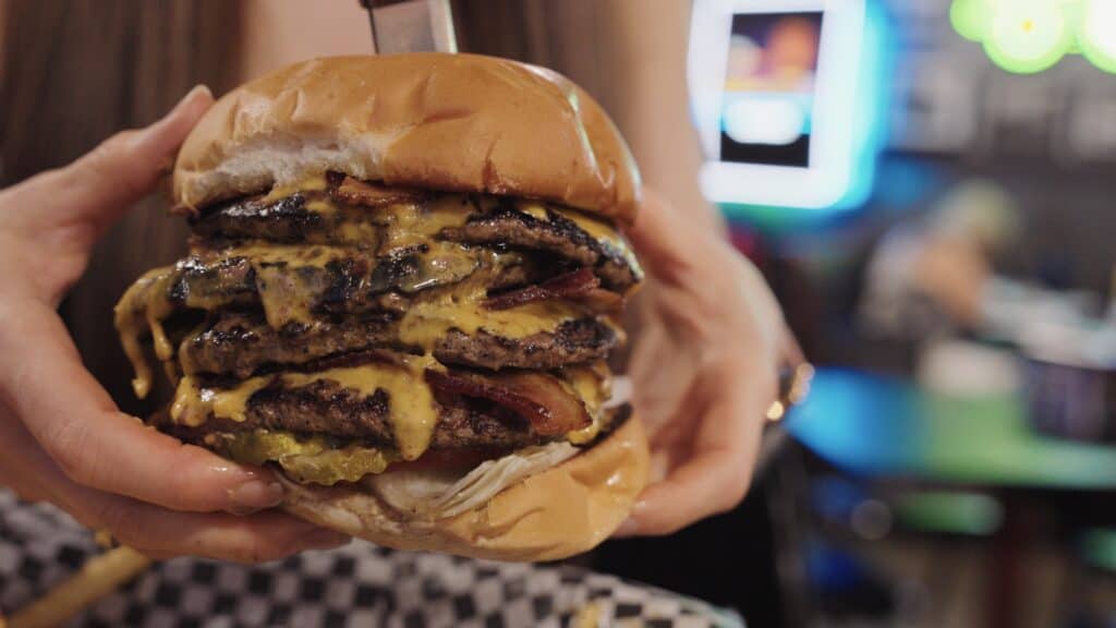 Hands holding an oversized triple cheeseburger stacked with grilled beef patties, melted cheese, bacon, pickles, and onions on a soft bun. Massive burgers like this are a memorable find at the best restaurants in Fort Dodge Iowa.