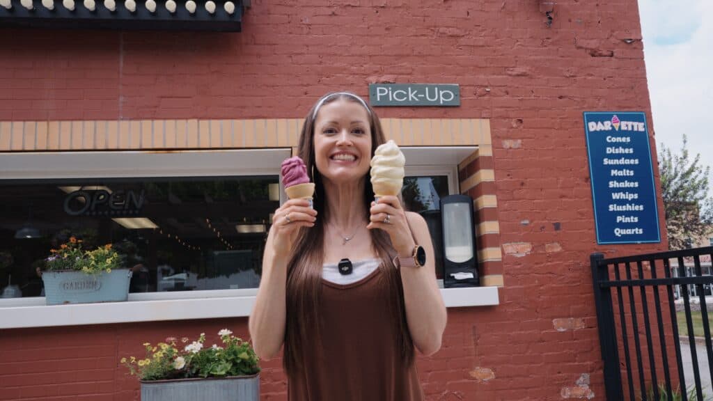Woman smiling outside a classic ice cream shop while holding a purple scoop in one cone and vanilla soft serve in another. Local dessert stops are part of the diverse dining scene at the best restaurants in Fort Dodge Iowa.