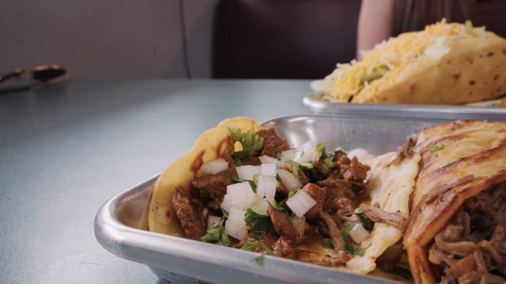 Soft corn tacos filled with seasoned beef, diced onion, and fresh cilantro served on a metal tray with a stuffed burrito in the background. Authentic Mexican flavors are a highlight of the best restaurants in Fort Dodge Iowa.