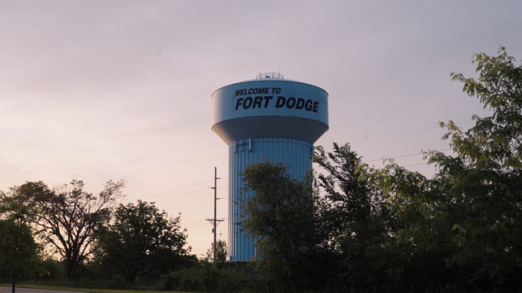Blue water tower displaying the words "WELCOME TO FORT DODGE" rising above trees at sunset. The landmark welcomes visitors exploring the best restaurants in Fort Dodge Iowa.
